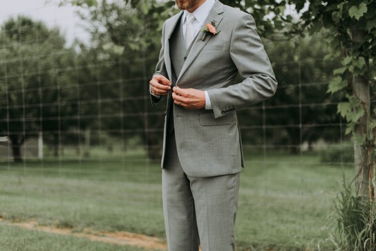 photo of a groom in a grey suit standing in a field buttoning his jacket