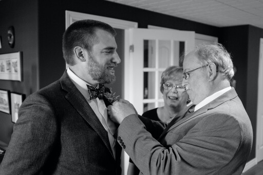 photo of a father pinning a flower on his son the groom while mom watches and smiles
