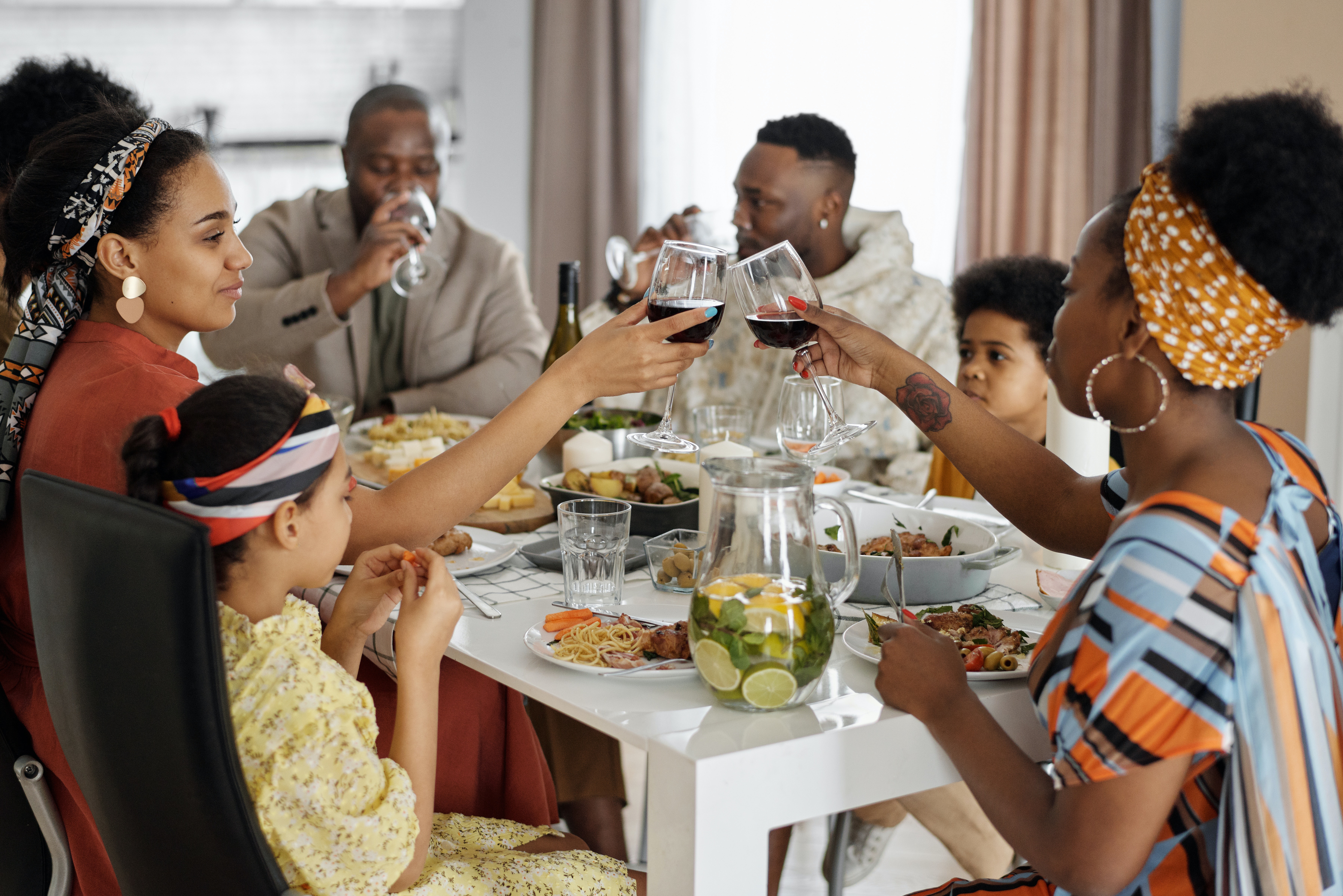 big family sits at a white dinner table with a diverse spread of food, toasting one another with red wine