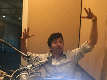 daniel post senning sitting at a radio microphone holding his hands up in a big gesture like he is holding an extra large beachball