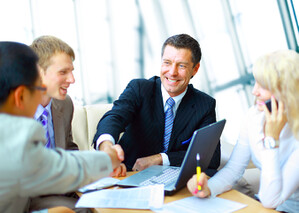 Man in a suit and tie smiles and shakes hands with a person sitting at a meetingroom table