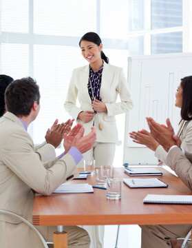 woman in a suit stands and smiles while her collegues offer applause arround a meeting table