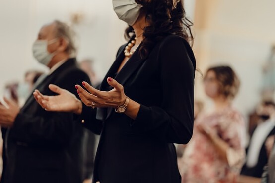 photo: woman at a service in black standing with her hands facing up wearing a mask