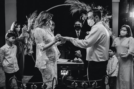 photo: bride and groom holding hands at the alter with priest and family wearing masks