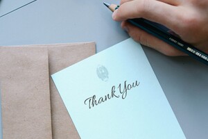 white thank you note atop beige envelope with a writer's hand holding a pencil above it