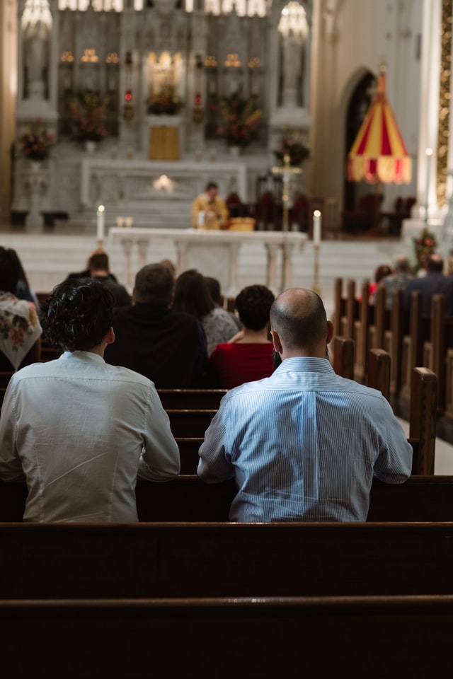 photo: two men kneel at a pew in a cathedral with priest at alter in background