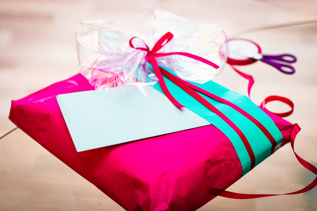 Flat square gift wrapped in hot pink tissue paper with an aqua accent and hot pink and white ribbons (pink ones are thin, white ones are wide) there's purple scissors in the background and a blank tag on the gift. it all sits on a soft brown paper background.