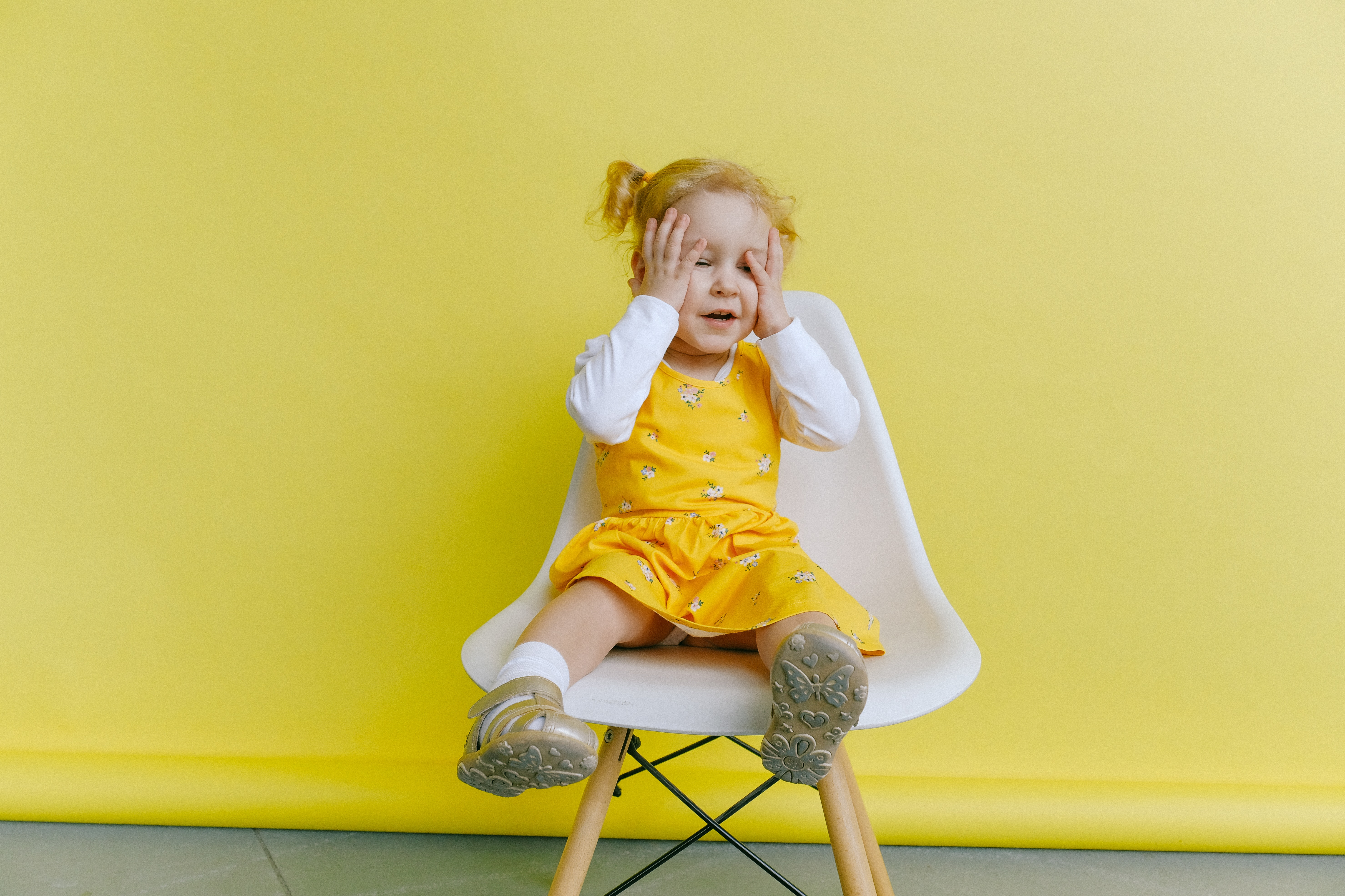 young child with her palms on her face sitting on a white chair with a yellow background