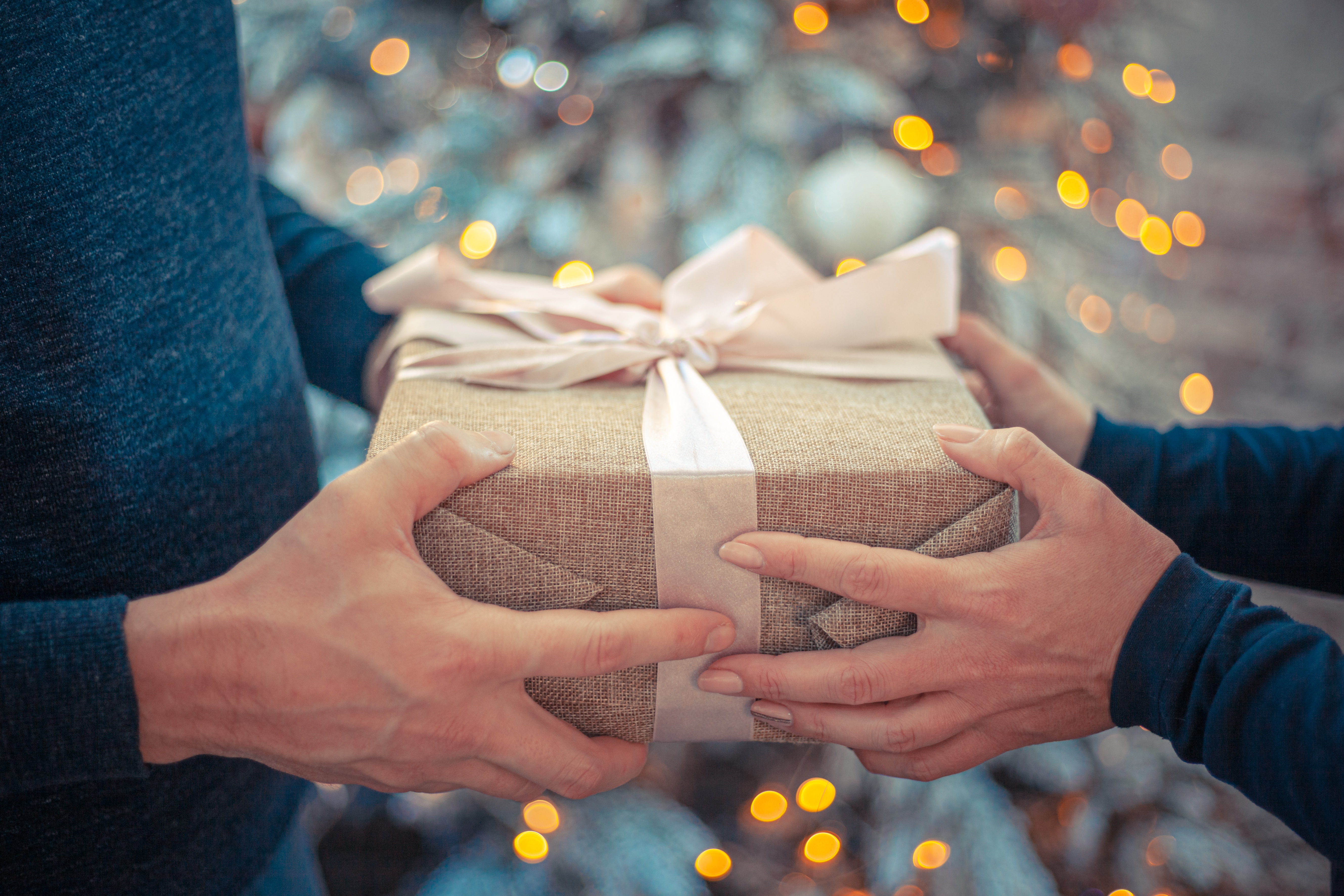 man and woman exchanging a wrapped gift with a decorated christmas tree in the background