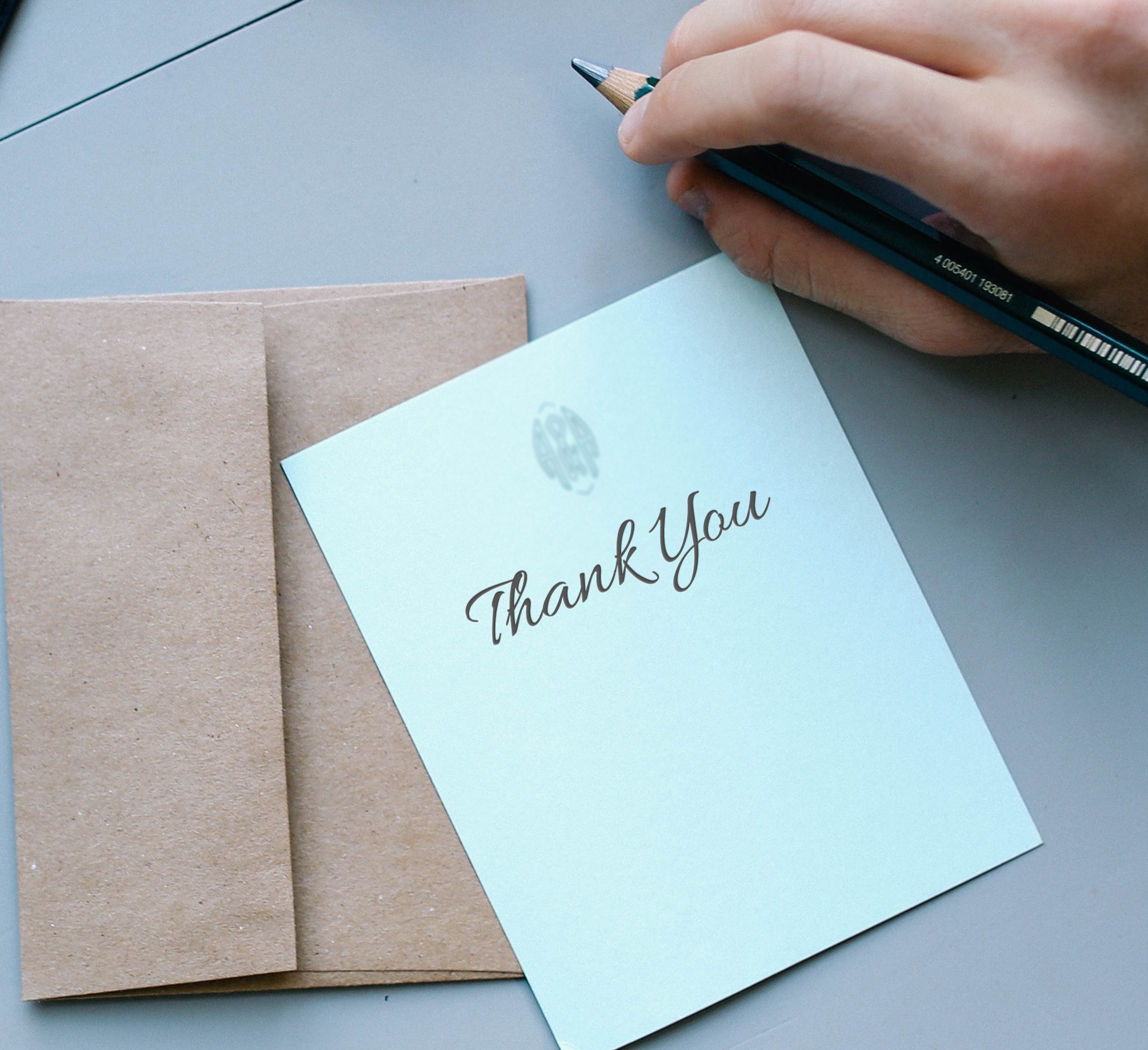 white thank you note atop beige envelope with a writer's hand holding a pencil above it