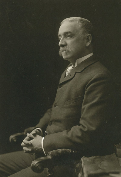 Black and white photo of EMily Post's father Bruce Price sitting with hands clasped in lap wearing three button suit.