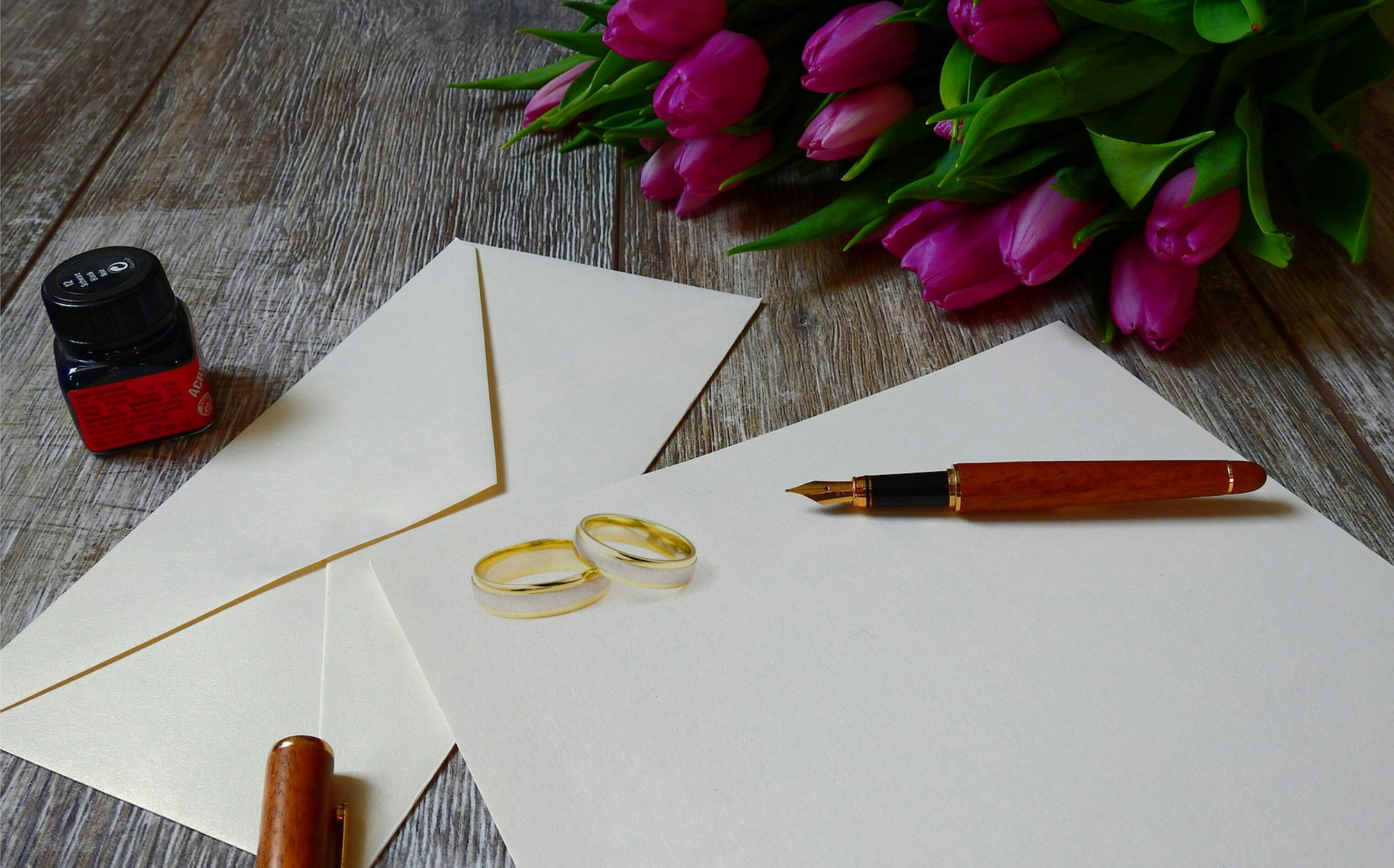 wedding invitations on a wooden table with a fountain pen, jar of ink, and wedding bands on top of them